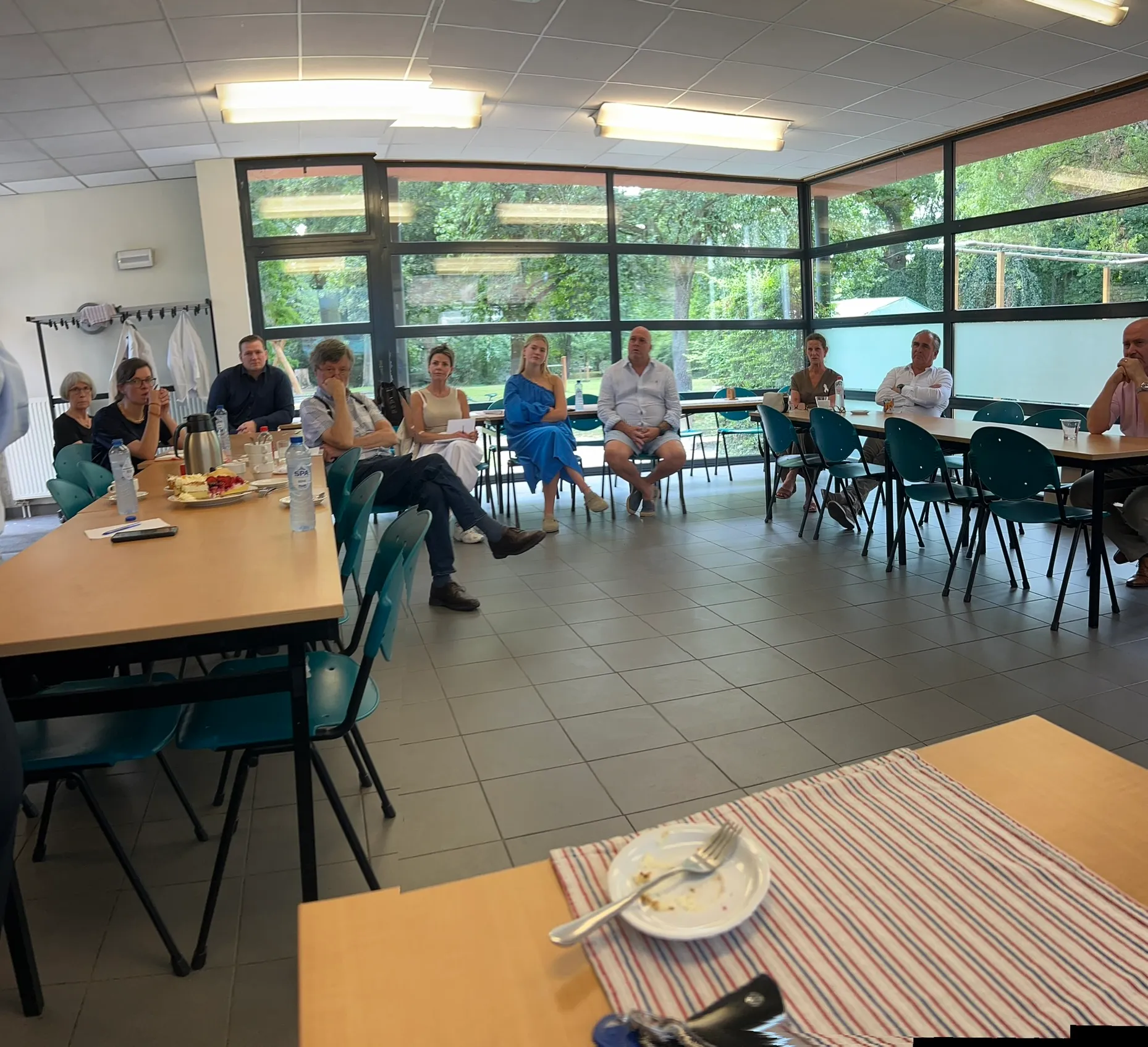 Een groep luistert in een grote kring tijdens een bijeenkomst, met koffie en taart op tafel